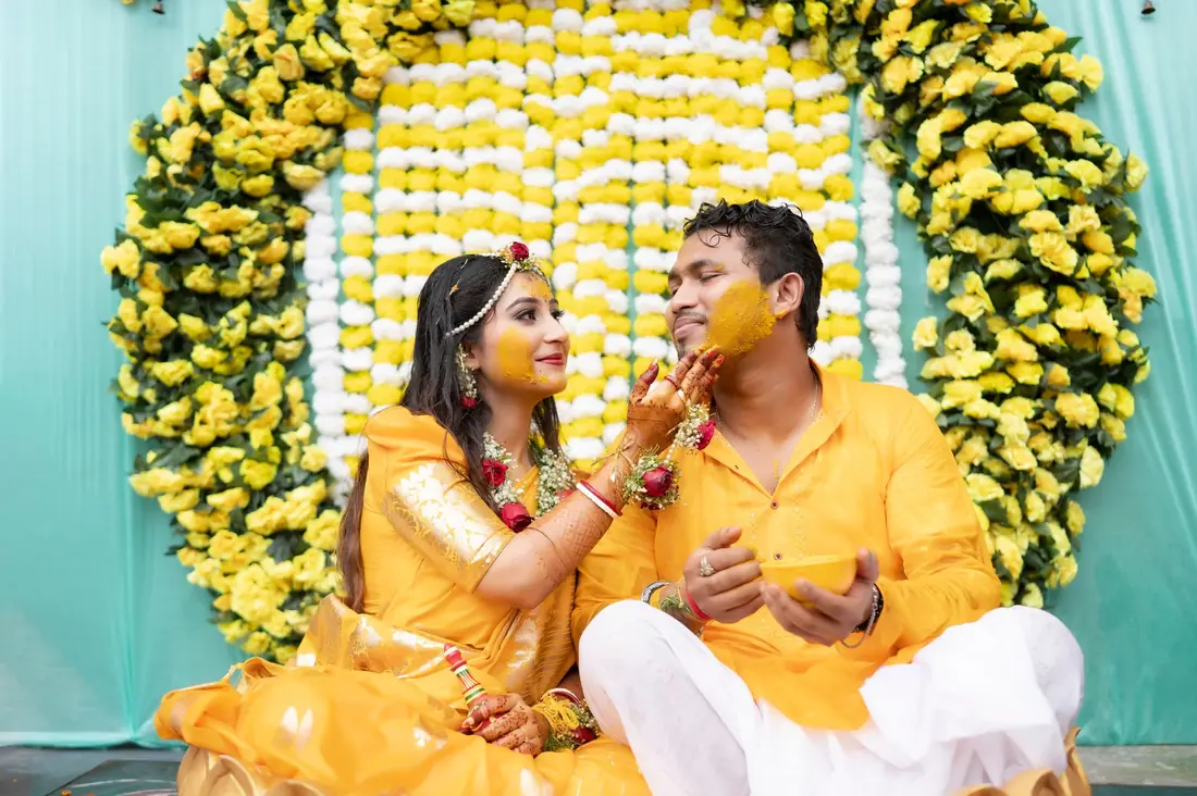 Couple at their haldi, the bride putting turmeric on the groom's cheek in front of a yellow and white flower wall